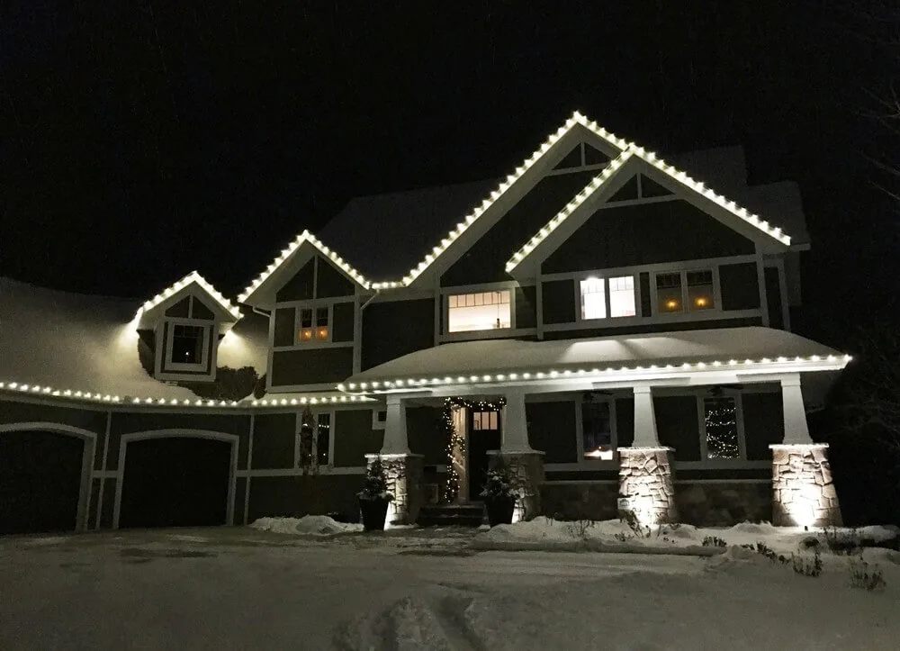 Two-story craftsman with warm white roofline lights along every gable and wrapped porch columns, snow on the ground