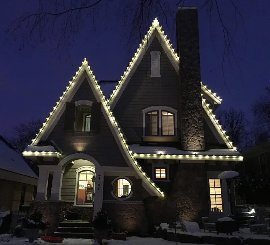 Gray two-story house with steep gables outlined in warm white roofline lights beside a brick chimney