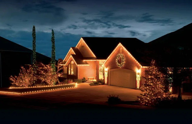 Stucco home at dusk with warm white roofline lights, a lit wreath above the garage and uplit shrubs