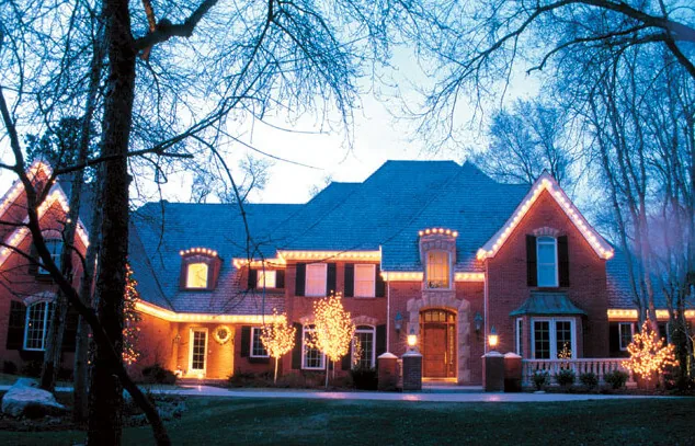 Large brick home at blue dusk with warm roofline lights and several wrapped trees, framed by bare branches