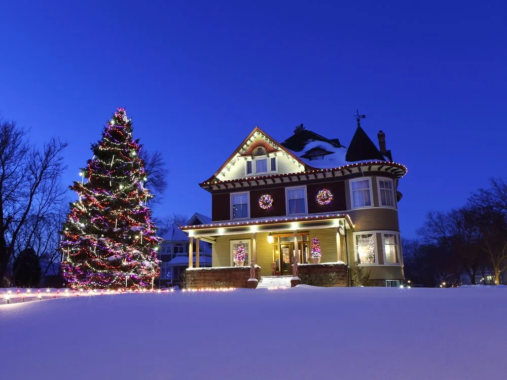 Victorian home in deep snow beside a tall evergreen wrapped in multicolor lights, warm porch lighting behind
