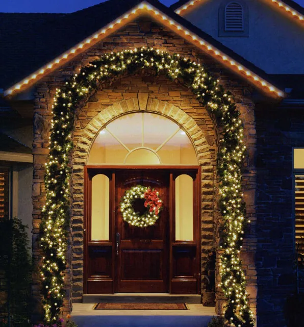Arched stone entry framed by a lit garland surround and a wreath, with warm white lights along the eave